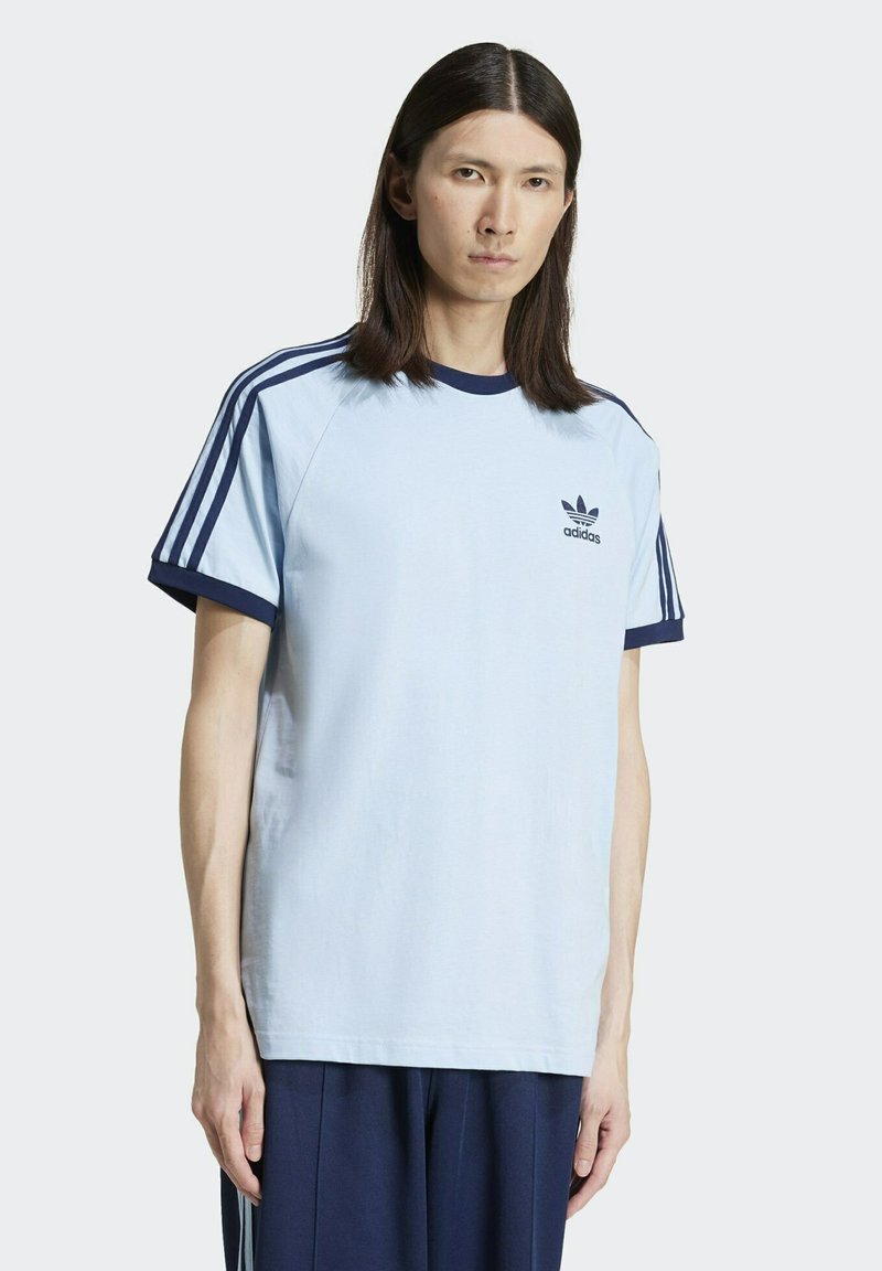 Camiseta de algodón azul claro con cuello y mangas acanalados en azul marino. Presenta tres rayas en azul marino en los hombros y un logo de Adidas en el pecho.