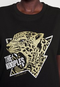 Camiseta de algodón negra con un gráfico de leopardo dorado y el texto "THE KOOPLES" debajo, además de un diseño geométrico blanco alrededor del gráfico.
