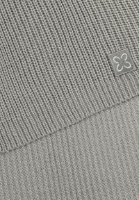 Helles graues gestricktes Material mit einer gerippten Textur. Ein quadratisches Stoffetikett mit einem dekorativen Design ist in einer Ecke sichtbar.