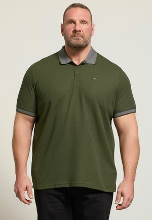 Muskuløs mand med skæg iført en olivengrøn polo med grå krave og ærmekanter, stående mod en ensfarvet lys baggrund.