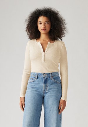T-shirt à manches longues en beige clair avec un col henley à boutons. Assorti à un jean bleu à taille haute et jambes larges. Texture lisse.