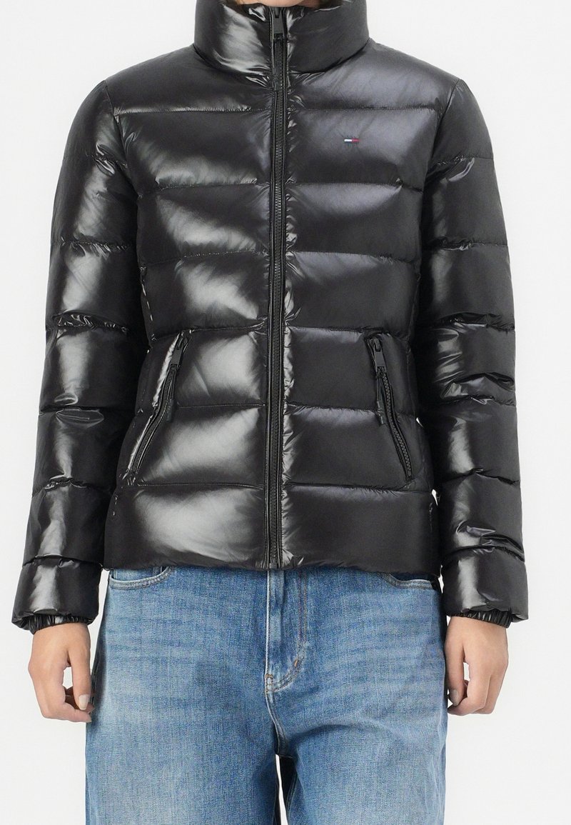 Personne portant une doudoune noire brillante avec un col haut et des poches zippées, associée à un jean bleu sur un fond uni.