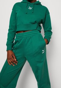 Sweat à capuche court vert avec un logo Puma blanc, associé à un pantalon de survêtement vert assorti avec une ceinture élastique et des coutures latérales.