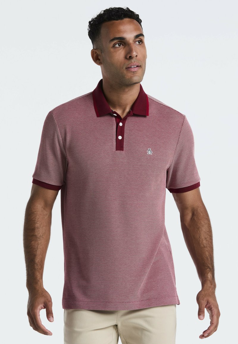 Un homme portant un polo bordeaux à manches courtes avec un col rouge foncé et un pantalon beige, regardant légèrement vers le haut et vers sa gauche.