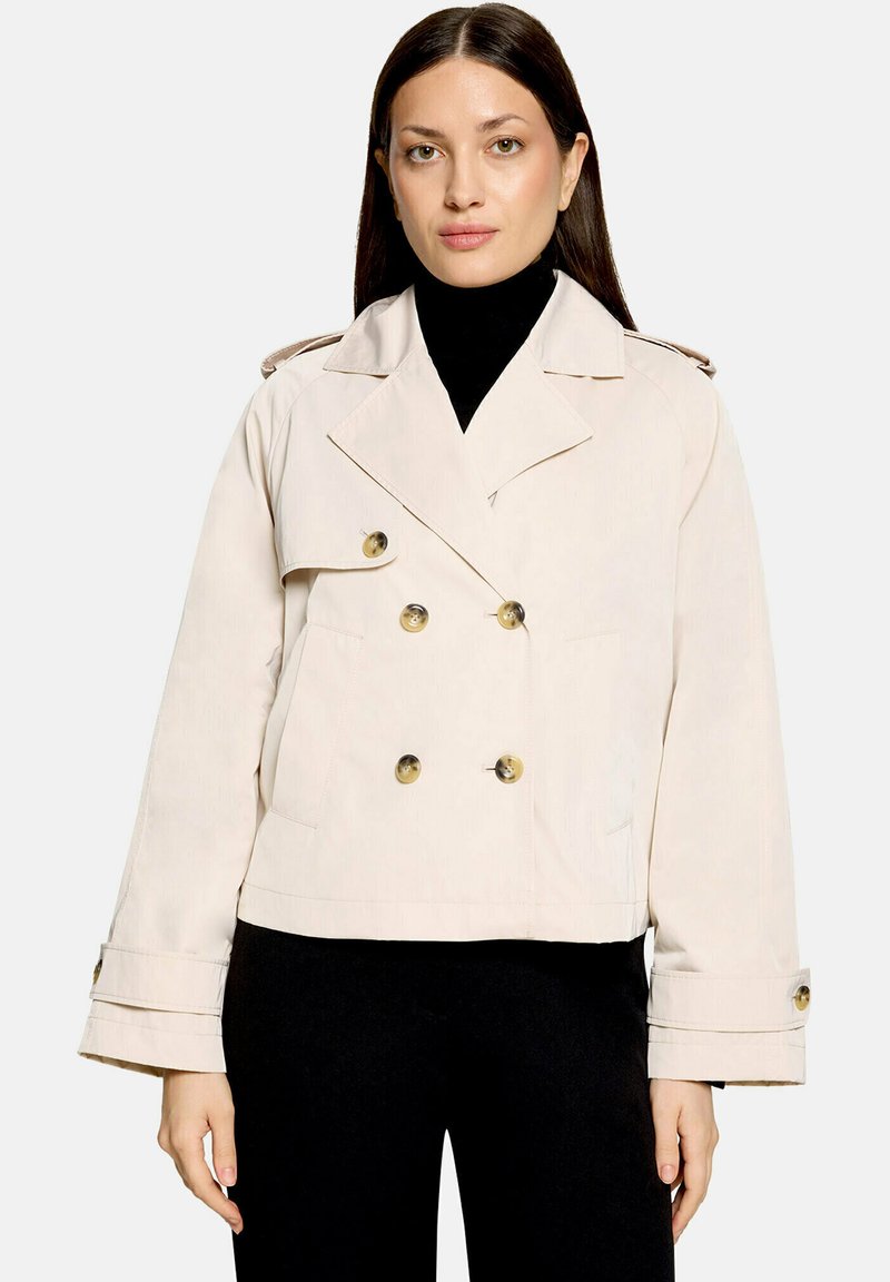 Femme aux cheveux longs et raides, vêtue d'une veste croisée crème par-dessus un col roulé noir et un pantalon noir, debout devant un fond uni.