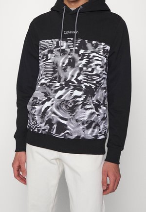 Mand iført sort hættetrøje med abstrakt sort-hvid mønstret frontpanel og hvidt Calvin Klein-logo, kombineret med hvide bukser.