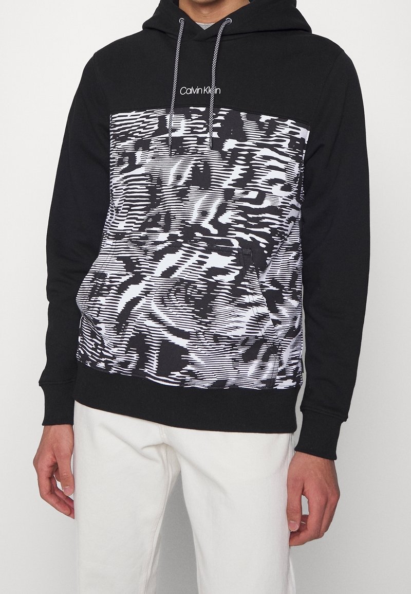 Homme portant un sweat à capuche noir avec un panneau avant à motif abstrait noir et blanc et un logo Calvin Klein blanc, associé à un pantalon blanc.