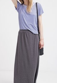 Gestreiftes blau-weißes T-Shirt mit kurzen Ärmeln, am Bund geknotet, kombiniert mit einem langen grauen Rock mit elastischem Bund. Dazu gehört eine schwarze Schultertasche.