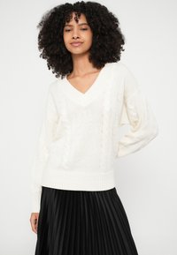 Maglione color crema con scollo a V, motivi testurizzati e polsini a costine, abbinato a una gonna nera plissettata, creando un look stratificato.