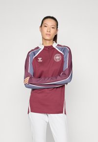 Bordeaux langærmet sportstop med halvlukning, hvid krave og blå striber. Har et rundt logo på brystet.