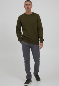 Oliven grønn genser med rund hals, kombinert med lys grå bukser og svarte joggesko. Jevn tekstur med minimal dekorasjon.