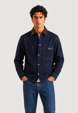 Mand med krøllet mørkt hår iført en marineblå jakke med knapper og brun krave samt blå jeans, hænderne i lommerne, neutral baggrund.