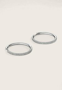 Silberne Creolen mit einem dünnen Band, verziert mit einer Reihe klarer Kristalle entlang des äußeren Randes. Glatte Metalloberfläche.