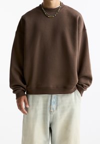 Brun oversized sweatshirt med ribstrikkede manchetter og kant, lavet af blødt materiale. Båret over en hvid skjorte og kombineret med lyseblå denimshorts.
