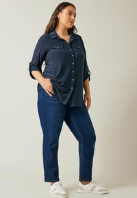 Camisa de botones azul marino con dos bolsillos en el pecho, mangas enrolladas y un corte relajado, combinada con jeans de mezclilla oscura y zapatillas blancas.