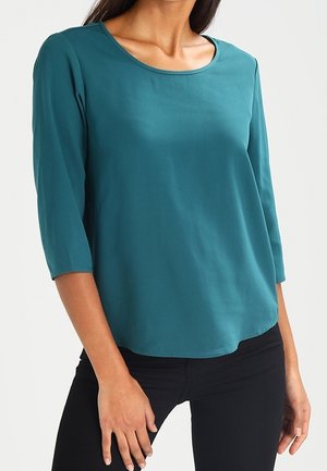 Femme portant un haut bleu sarcelle à manches longues et décolleté bateau, avec un pantalon noir, se tenant contre un fond clair.