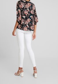 Woman wearing white skinny pants, white heeled sandals, and a black floral blouse with pink and white flowers, standing against a plain background.