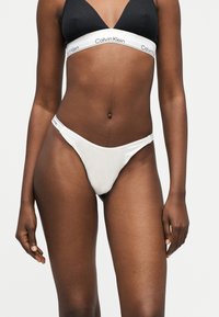 Γυναίκα που φοράει μαύρο μπραλέτα και λευκά εσώρουχα Calvin Klein με λογότυπο στις ελαστικές ταινίες, στέκεται μπροστά σε απλό φωτεινό φόντο.