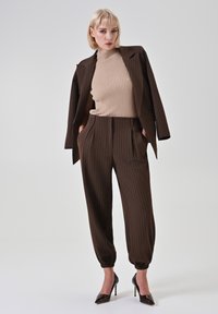 Blazer marrone a righe sottili indossato sopra un maglione beige a coste e pantaloni coordinati con polsini elasticizzati, abbinati a scarpe nere con tacchi alti.