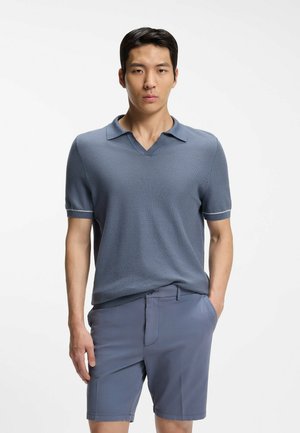 Giovane uomo che indossa una polo a maniche corte blu tenue e pantaloncini abbinati, in piedi con una mano in tasca su sfondo bianco semplice.