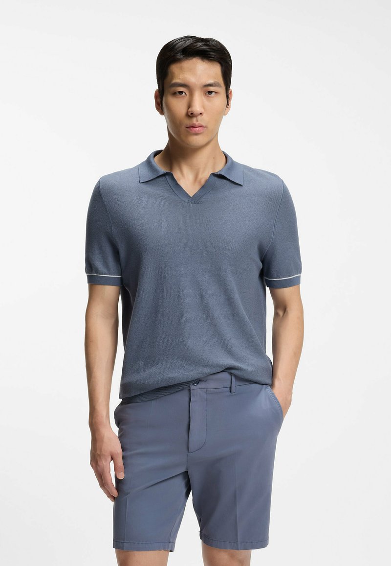 Jeune homme portant un polo à manches courtes bleu doux et un short assorti, debout avec une main dans la poche sur un fond blanc uni.