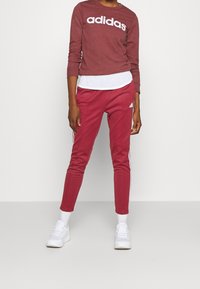Personne portant un sweat Adidas bordeaux et un pantalon de survêtement assorti avec des baskets blanches, debout devant un fond blanc uni.