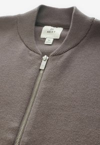Brown knitted cardigan featuring a ribbed collar, full zip closure, and metallic zipper detail. Contains a label with brand and size information.