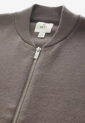 Cardigan tricotat de culoare maro, cu guler ribit, închidere cu fermoar complet și detalii metalice la fermoar. Conține o etichetă cu informații despre marcă și mărime.