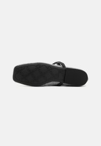 Semelle plate noire de chaussure avec motif géométrique texturé et talon lisse, vue du dessous sur un fond uni.