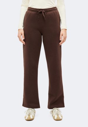 Personne portant un pantalon de survêtement marron avec cordon de serrage et des baskets blanches, debout, les mains détendues le long du corps, devant un fond uni.