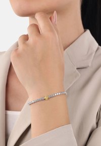 Primer plano de una muñeca con una pulsera de tenis de plata con una gema amarilla en forma de estrella, mano descansando cerca del cuello, blazer beige visible.