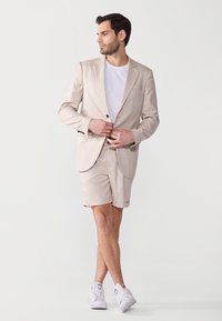 Blazer beige à un bouton, short beige assorti et t-shirt blanc en dessous. Le modèle porte des sneakers blanches. Tissu lisse et coupe ajustée.