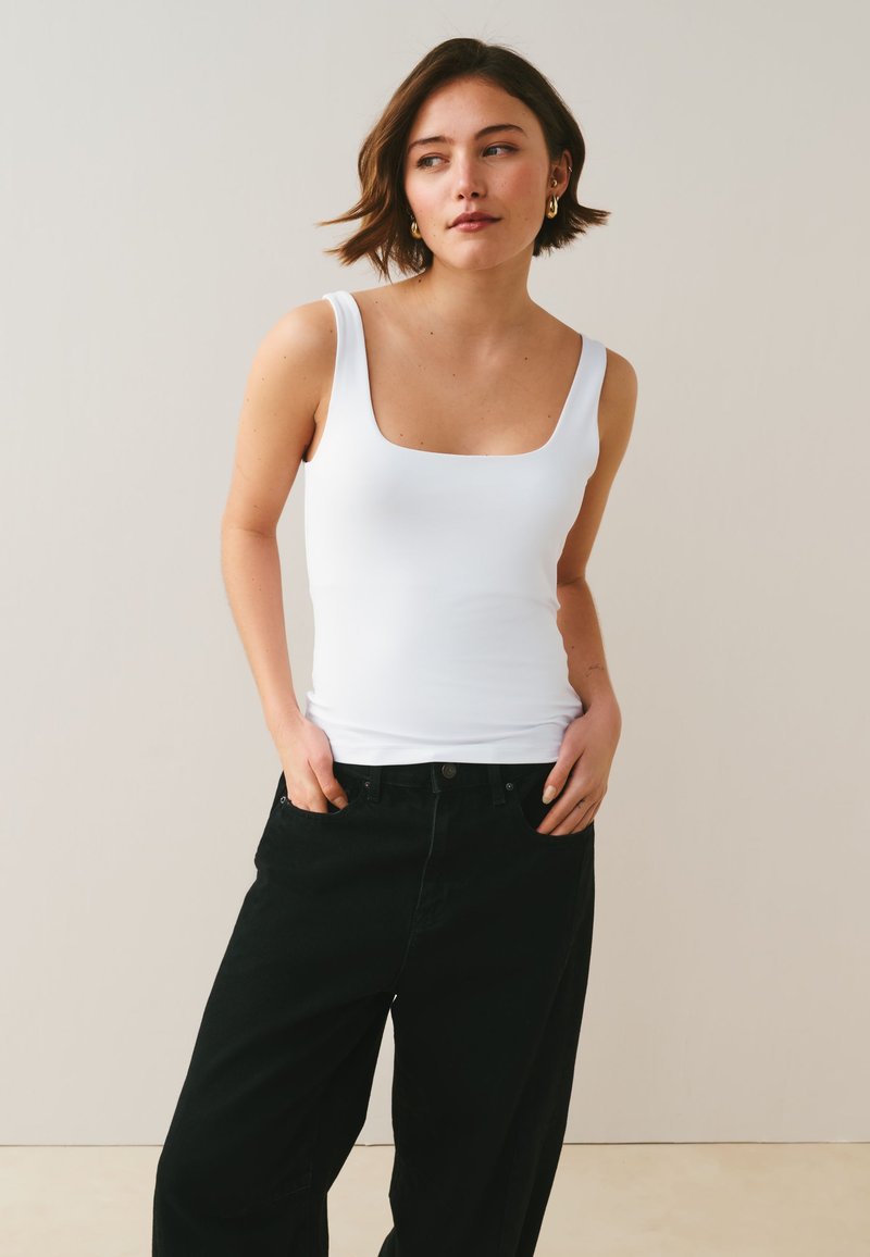 Woman with short brown hair wearing gold hoop earrings, a white tank top, and black pants stands with hands in pockets looking sideways.