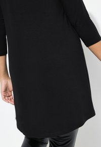 Schwarze Tunika mit Dreiviertelärmeln, glatter Textur, abgerundetem Saum und seitlichen Schlitzen, kombiniert mit glänzenden schwarzen Leggings.