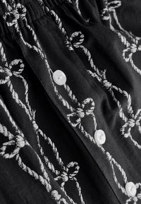 Fekete szövet fehér kötéldíszítésmintával. Texturált felület, redős derékpánttal és jól látható fehér műanyag gombokkal.