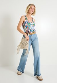 Top sin mangas de colores y estampados, jeans acampanados de azul claro, cinturón marrón, bolsa de mimbre beige con flecos y zapatillas claras.