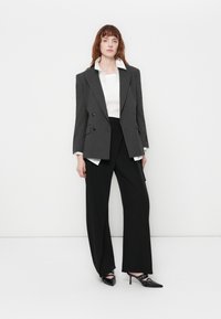 Mujer con cabello rojo vestida con una chaqueta gris de rayas, camisa blanca, pantalones de pierna ancha negros y tacones negros de punta, de pie contra un fondo blanco.