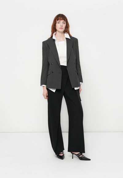 Femme aux cheveux roux portant un blazer gris à rayures, une chemise blanche, un pantalon noir à jambes larges et des escarpins noirs à bout pointu, se tenant devant un fond blanc.