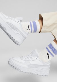 Adidași albi cu panouri texturate și talpă din cauciuc, asociați cu șosete albe cu dungi albastre și mov și un logo negru.