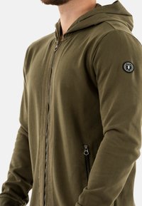 Veste à capuche zippée en vert olive fabriquée dans un tissu doux, avec un zip avant, une poche latérale et un patch logo sur la manche gauche.