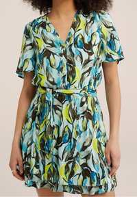 Kvinna i kortärmad klänning med V-ringning, blå, grön och brun abstrakt bladmönster, knappstängning fram och matchande bälte av samma tyg.