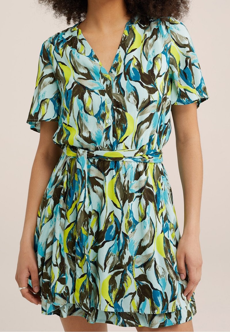 Kvinna i kortärmad klänning med V-ringning, blå, grön och brun abstrakt bladmönster, knappstängning fram och matchande bälte av samma tyg.