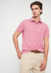 Šviesiai rožinė polo marškinėliai su klasikinio tipo apykakle, dviem mygtukais ir mažo logotipo ant krūtinės; derinami su šviesiai smėlinėmis kelnėmis ir rudu diržu.