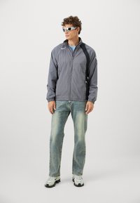 Grå vindjakke med elastiske manchetter og et sort sidepanel, parret med lyseblå jeans og hvide sneakers. Modellen bærer refleksbriller.