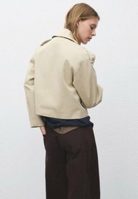 Beige kort jacka med slagkrage och axeldetaljer, bärs över en marinblå skjorta och mörkbruna byxor, mot en enkel bakgrund.
