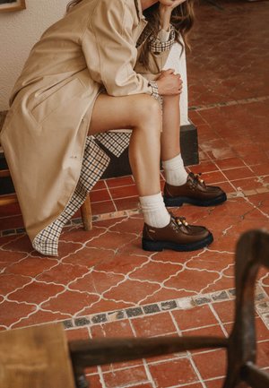 Femme assise sur une chaise, une jambe croisée, portant un manteau beige, une robe à carreaux, des chaussettes blanches et des chaussures en cuir marron à lacets.