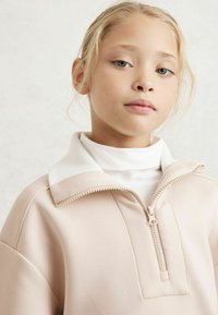 Felpa zipata in morbido tessuto beige, con colletto a coste bianco e zip frontale a metà. Trama liscia con dettagli di design minimalisti.