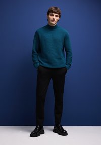 Pull côtelé teal avec un col montant, associé à un pantalon noir et des chaussures noires épaisses, sur fond bleu.