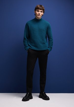 Maglione teal a coste con colletto alto, abbinato a pantaloni neri e scarpe nere chunky, su uno sfondo blu.
