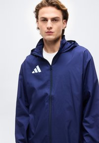 Νεαρός άνδρας που φοράει μπλε σκούρο φούτερ adidas με φερμουάρ και κουκούλα, λευκό πουκάμισο από μέσα, μπροστά σε ένα απλό ανοιχτό φόντο.